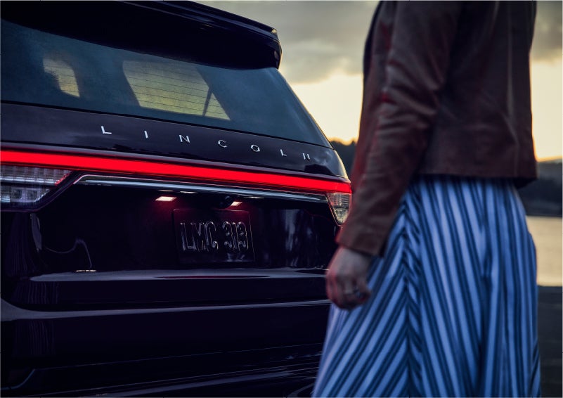 A person is shown near the rear of a 2023 Lincoln Aviator® SUV as the Lincoln Embrace illuminates the rear lights | Varsity Lincoln in Novi MI